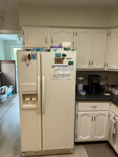 A cream refrigerator covered in magnets stands beside white cabinetry in a kitchen awaiting renovation.