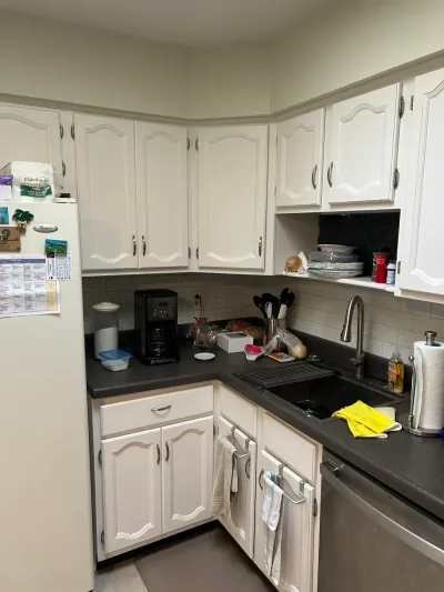A cluttered kitchen corner with white cabinets, dark countertops, and subway tile backsplash before renovation.