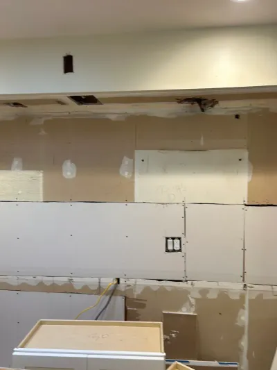 Kitchen wall mid-renovation with unfinished drywall seams, exposed wiring, and a partially installed white cabinet.
