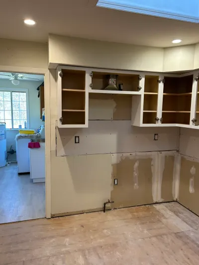 Kitchen mid-renovation with empty upper cabinets and lower cabinets removed down to bare drywall and subfloor.