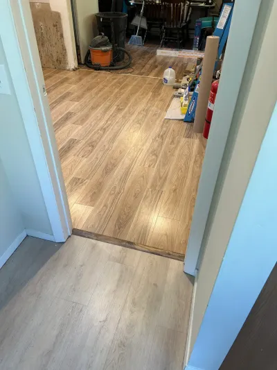 A doorway shows two different wood-look flooring installations meeting at a transition strip, with tools and supplies scatter