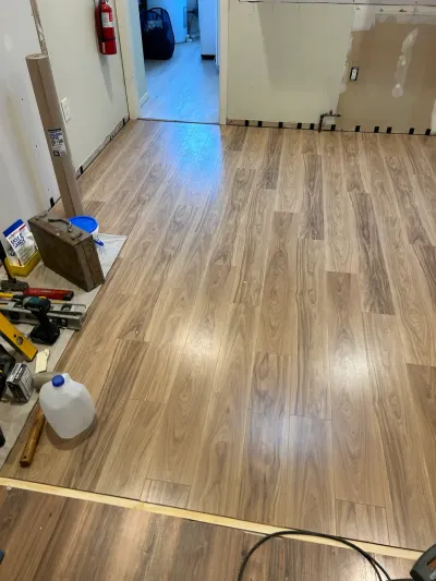 A basement mid-renovation with new plank flooring installed and an exposed wall section revealing studs and plumbing.