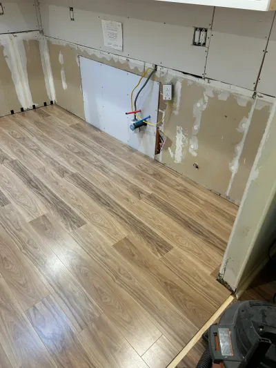 Kitchen mid-renovation with new plank flooring installed and walls stripped to bare drywall with exposed plumbing.