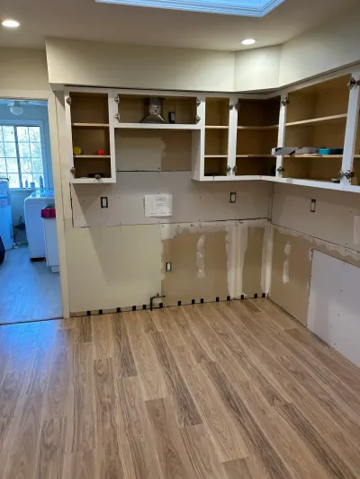 A kitchen mid-renovation with new flooring installed, upper cabinets hung but empty, and bare walls awaiting lower cabinetry.