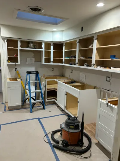Kitchen under construction with white cabinet boxes installed, doors removed, and a shop vacuum and ladders on the floor.
