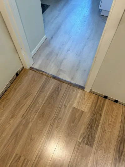 Close-up of a doorway where warm brown laminate flooring meets gray wood-look flooring at a worn transition strip.