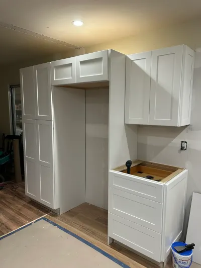 White shaker cabinets being installed in a kitchen, with an unfinished countertop base and taped-off flooring.