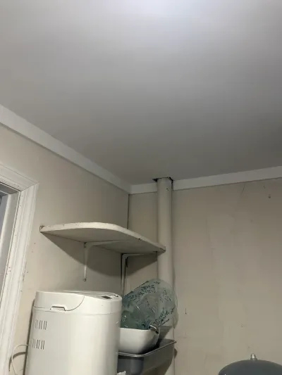 Basement corner with a pipe running through the ceiling, a wall shelf, and household items below.