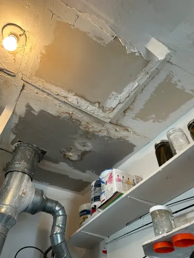 Damaged, water-stained basement ceiling with peeling patch work near ductwork and storage shelves.