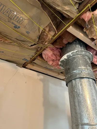 Close-up of exposed basement ceiling joists with pink insulation and a metal vent pipe passing through.
