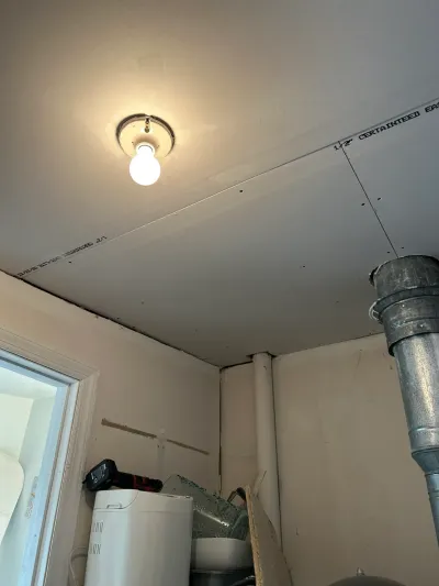 Unfinished ceiling and wall drywall in a utility room with exposed ductwork and a bare light bulb.
