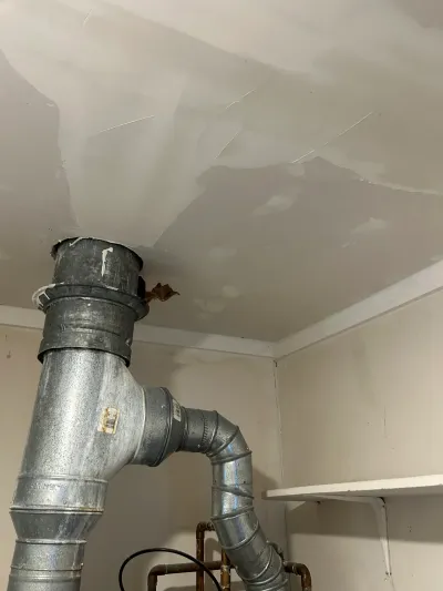 Basement ceiling with a patched drywall repair around a large galvanized ductwork elbow.