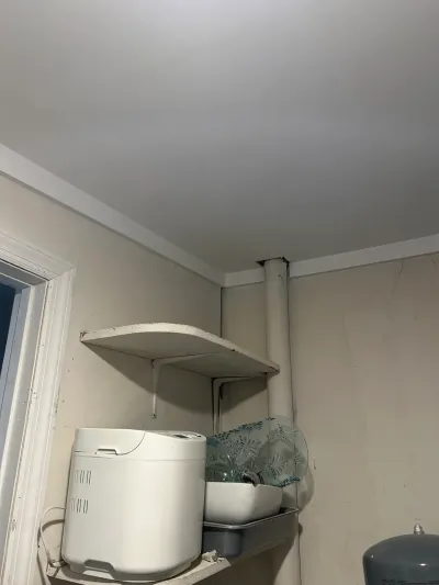 Corner of a cluttered utility room with a shelf holding a bread maker and an exposed pipe along the wall.