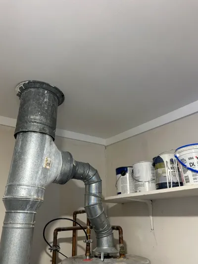 Galvanized flue pipe from a water heater bends through the ceiling near a shelf stacked with paint cans.