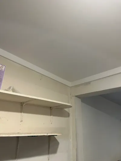 Corner of a storage closet with worn shelving and crown molding, showing an open doorway to another room.