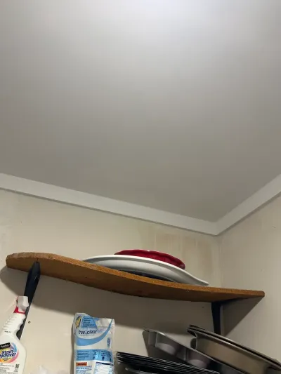 Close-up of a stained wall corner near the ceiling above a shelf holding bakeware and cleaning supplies.