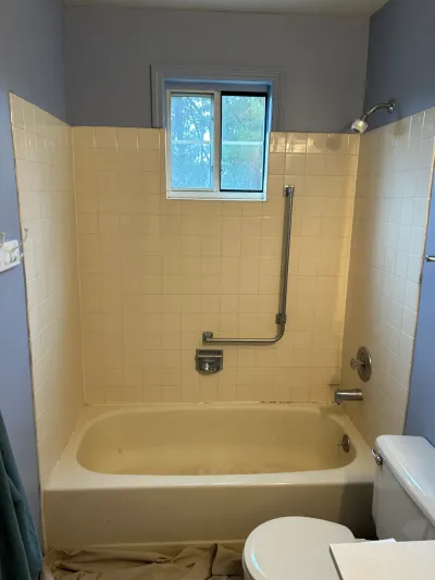 Dated bathroom tub and tile shower surround before renovation, with a window and drop cloth on the floor.