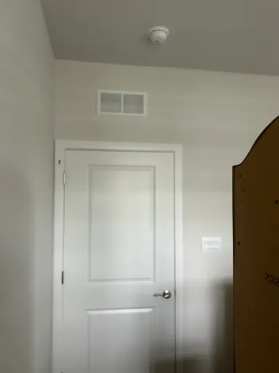 A finished hallway with a white paneled door, ceiling vent, smoke detector, and thermostat.