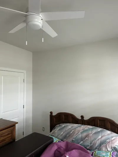 A bedroom with a white ceiling fan, light gray walls, and a wooden headboard with patterned bedding.