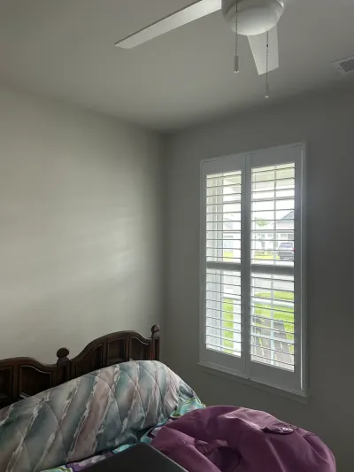 A bedroom corner with a wood headboard, patterned bedspread, and a window with white plantation shutters.