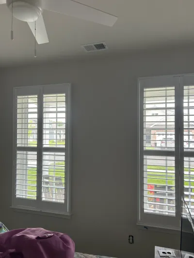 Bedroom corner with two large windows fitted with white plantation shutters and a ceiling fan.