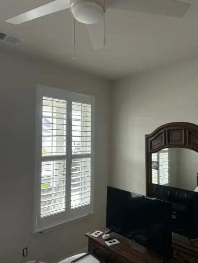 Bedroom corner with plantation shutters, a ceiling fan, and a wood dresser topped with a mirror and TV.