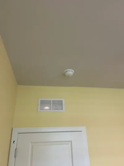 Ceiling corner with a smoke detector, air vent, and yellow wall above a white door with painter's tape.