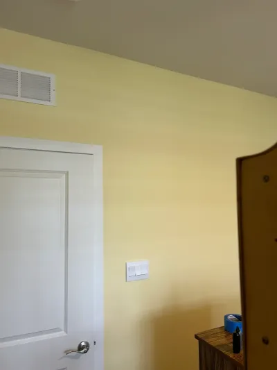 Pale yellow bedroom wall with a white paneled door, air vent, and light switch plate.