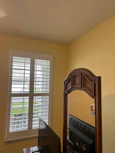 A yellow-walled bedroom corner with a shuttered window and a large wooden arched mirror reflecting a TV.