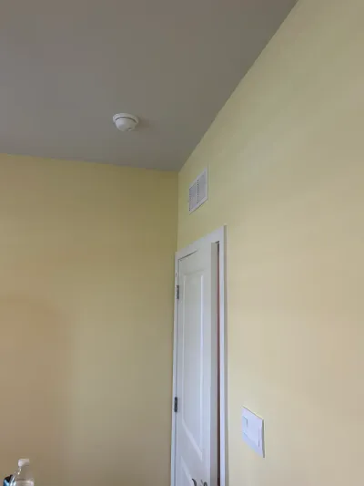 Corner of a room with pale yellow walls, a gray ceiling, a smoke detector, and an HVAC vent above a white door.