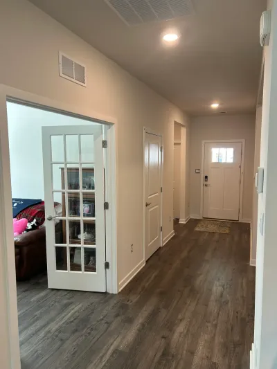 A finished hallway with dark wood-look flooring leads to a white front door, with a glass-paned door opening into a living ro