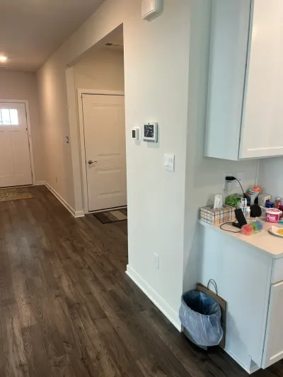 A hallway with dark wood-look flooring leads to a front door, with white kitchen cabinets visible nearby.