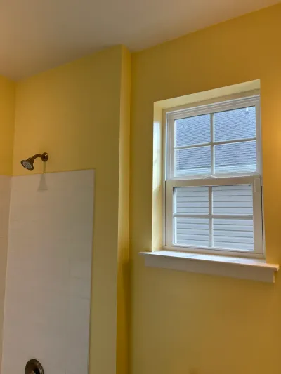 Bathroom corner with white subway tile shower, bronze shower head, yellow walls and a white double-hung window.