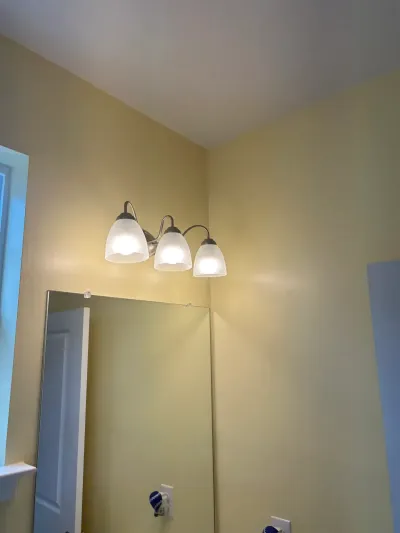 Three-light brushed nickel vanity fixture above a frameless mirror on a pale yellow bathroom wall.
