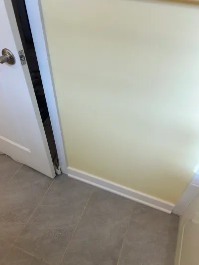 Corner detail of cream wall, white baseboard, gray tile floor and a white door with brushed nickel knob.