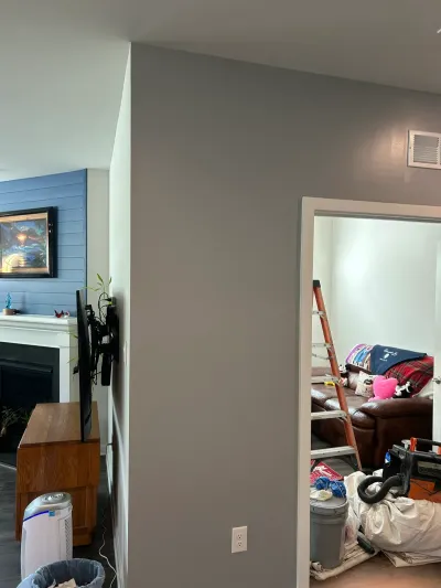 Gray living room wall beside a doorway into a room staged with a ladder, drop cloths and tools.