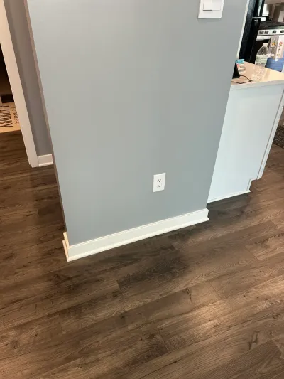 Corner of a blue-gray wall with white baseboard meeting dark wood-look vinyl plank flooring near a kitchen.
