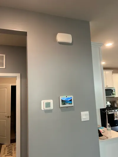 Gray-blue hallway wall with thermostat and smart home touchscreen, kitchen with white cabinets beyond.