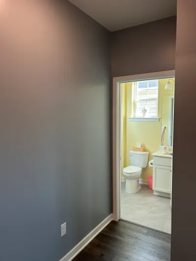 Hallway with gray-blue walls looking through a doorway into a yellow bathroom with toilet and white vanity.