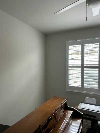 Greige office corner with white plantation shutters, ceiling fan, and a wooden desk holding a printer.