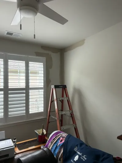 Bedroom mid-repair with patched primer spots on walls, a step ladder, paint bucket, and shuttered window.