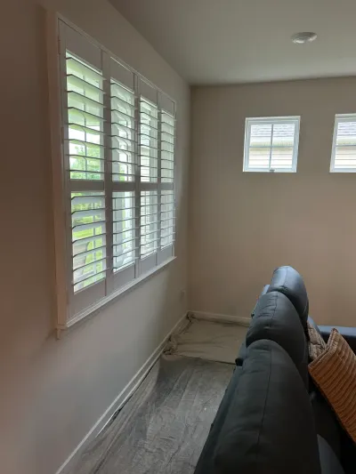 Freshly painted taupe living room corner with white plantation shutters, drop cloths on floor, navy sofa.