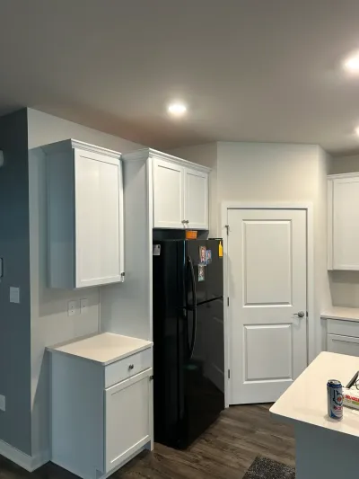 White shaker kitchen cabinets and quartz counters around a black refrigerator, dark vinyl plank floor.