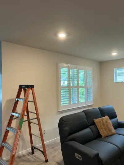Freshly painted living room with plantation shutters, recessed lights, step ladder and gray leather sofa.