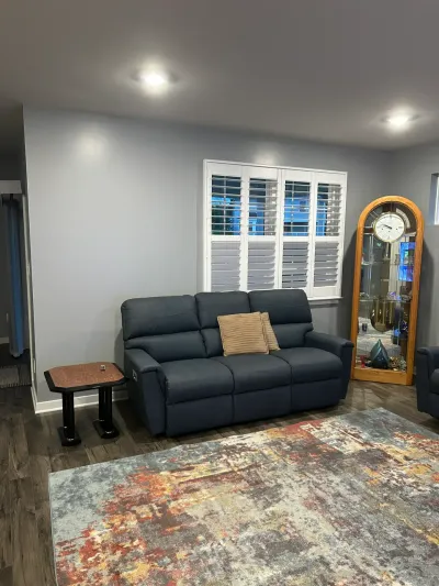Living room with navy reclining sofa under white plantation shutters, oak clock, and abstract area rug.
