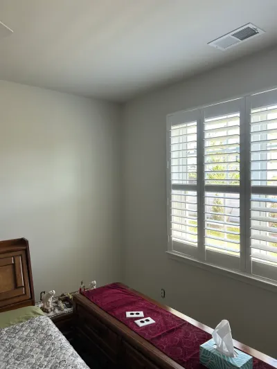 Bedroom corner with greige walls, white plantation shutters, and a dark wood dresser beneath the window.