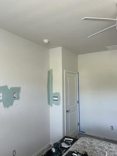 Bedroom corner mid-paint project with green test swatches on white walls, taped door, and tools on the floor.