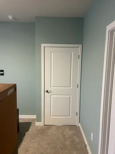 Bedroom corner with soft blue-green walls, white two-panel door, white trim and beige carpet.