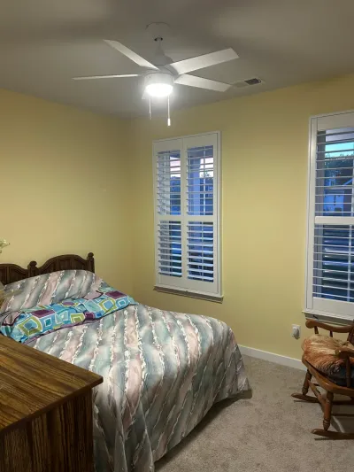 Yellow bedroom with white plantation shutters, beige carpet, ceiling fan, wood bed and rocking chair.