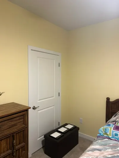 Bedroom corner with pale yellow walls, white two-panel door, beige carpet, wood dresser and storage ottoman.